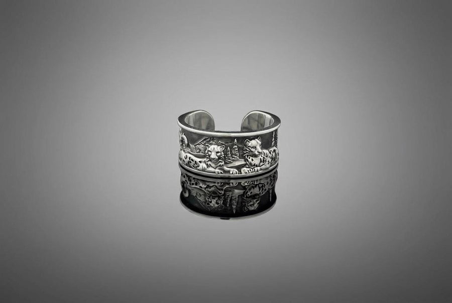 A close-up studio shot of a wide, open-back sterling silver cuff ring resting on a black reflective surface. The band features a detailed, three-dimensional bas-relief carving of a mountain landscape. In the foreground, two realistic snow leopards lie side by side among textured pine trees and rocky peaks. The 925 silver is heavily oxidized in the recesses to highlight the intricate fur spots, trees, and mountainous terrain against the polished r __etsy:7979063548