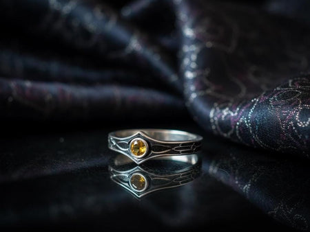 A cinematic close-up of a sterling silver ring with a distinct "V" shaped or "Lance Tip" contour. The focal point is a bezel-set, round gemstone with a deep amber-orange hue (Citrine or Madeira Citrine). The shoulders of the band feature horizontal grooves that taper toward the center, resembling the shaft of a spear or the fletching of an arrow. The metal is polished but features dark oxidation in the grooves for contrast.