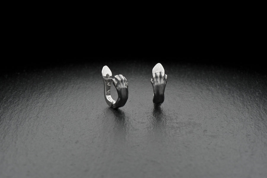 A close-up studio shot of a pair of sterling silver huggie hoop earrings resting on a dark slate surface. The front of each earring is masterfully sculpted to resemble a realistic cat paw, complete with distinct knuckles, claws, and soft paw pads. The earrings feature a hinged mechanism that allows the paw to wrap seamlessly around the earlobe. The metal is treated with heavy black oxidation in the recesses, contrasting beautifully with the brigh (etsy image id: 4850613175)