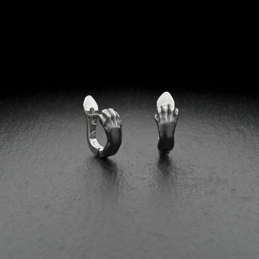 A close-up studio shot of a pair of sterling silver huggie hoop earrings resting on a dark slate surface. The front of each earring is masterfully sculpted to resemble a realistic cat paw, complete with distinct knuckles, claws, and soft paw pads. The earrings feature a hinged mechanism that allows the paw to wrap seamlessly around the earlobe. The metal is treated with heavy black oxidation in the recesses, contrasting beautifully with the brigh (etsy image id: 4850613175)