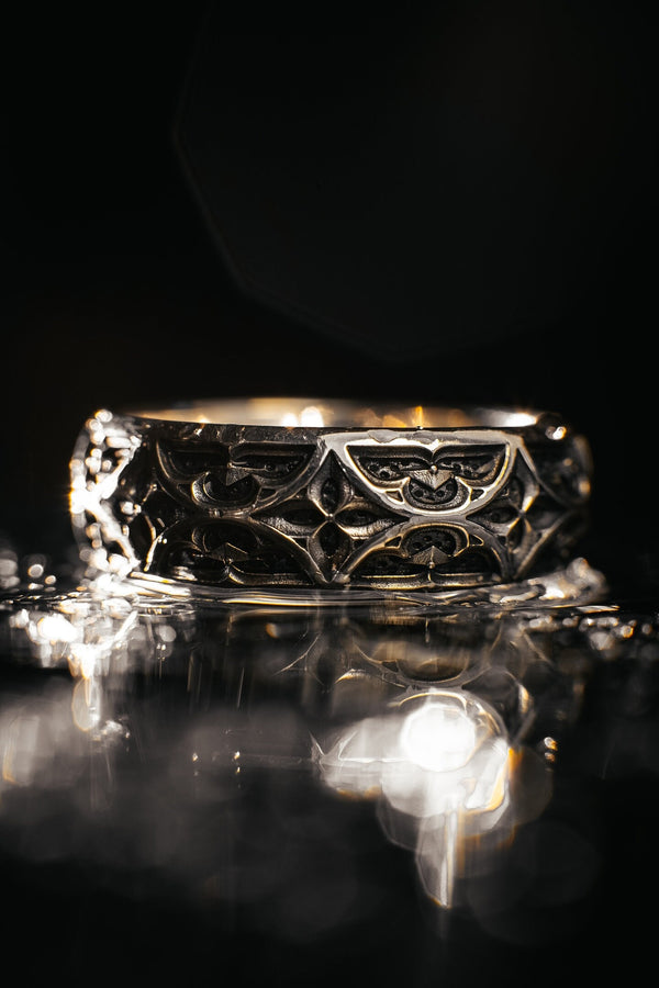 A high-resolution macro photograph of a matching sterling silver gothic wedding ring set resting on softly draped, luxurious white satin. The set features a wider masculine band and a slightly narrower feminine band. Both rings display a continuous engraved arcade of pointed medieval arches along the bottom edge, centering on a polished geometric rhombus frame that holds a brilliant round white gemstone. The polished silver beautifully contrasts  (etsy image id: 7928017042)
