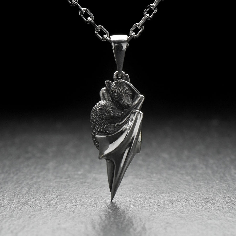 A detailed close-up studio shot of a sterling silver pendant shaped like two flying foxes or fruit bats. The bats are intimately huddled together, wrapped within the smooth, sweeping folds of their leathery wings. The metal is highly textured to show the fine details of their fur and faces. The silver is heavily oxidized in the recesses, creating a striking contrast against the polished highlights of the wings. (etsy image id: 5592925539)