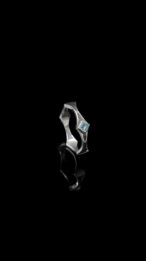 This Art Deco wedding ring set features a sterling silver engagement ring and matching band with bold geometric cutouts and a bright blue square gemstone. The clean lines, layered shapes, and sculptural silhouette create a modern yet vintage feel ideal for couples seeking unique, architecturally inspired bridal jewelry captured on a reflective surface for elegant presentation.
