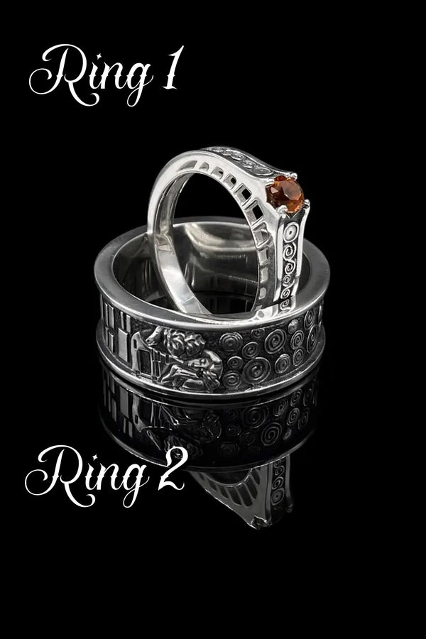 A cinematic studio shot of a matching two-piece ring set. The bottom ring is a wide silver band featuring a detailed relief carving of the couple from Gustav Klimt's "The Kiss." The top ring is a narrower engagement band that mimics the geometric patterns of the painting and holds a round, amber-colored gemstone. The background is a warm, golden haze, reflecting the "Golden Phase" of the original artwork.
