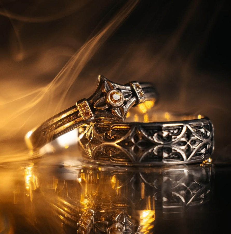 A high-resolution macro photograph of a matching sterling silver gothic wedding ring set resting on softly draped, luxurious white satin. The set features a wider masculine band and a slightly narrower feminine band. Both rings display a continuous engraved arcade of pointed medieval arches along the bottom edge, centering on a polished geometric rhombus frame that holds a brilliant round white gemstone. The polished silver beautifully contrasts  (etsy image id: 7928017042)