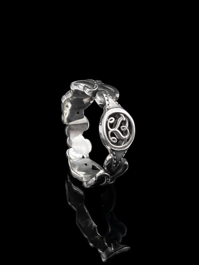 A studio shot of a matching two-piece sterling silver ring set on a black reflective surface. The standing ring is a signet style featuring an oval face with a raised Triskelion (triple spiral) motif. The ring lying flat is a wide band with an "openwork" or cut-out Celtic knot pattern. The silver is highly polished on the raised surfaces, contrasting with the dark shadows of the open areas.