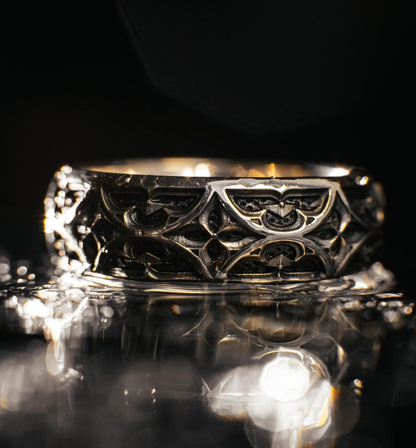 A high-resolution macro photograph of a matching sterling silver gothic wedding ring set resting on softly draped, luxurious white satin. The set features a wider masculine band and a slightly narrower feminine band. Both rings display a continuous engraved arcade of pointed medieval arches along the bottom edge, centering on a polished geometric rhombus frame that holds a brilliant round white gemstone. The polished silver beautifully contrasts  (etsy image id: 7928017042)