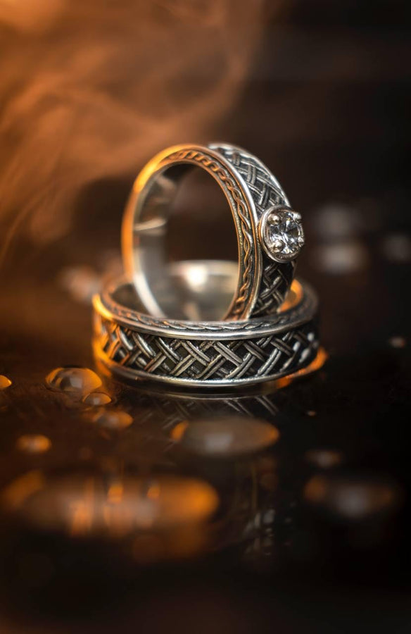 A warm, atmospheric shot of a matching wedding ring set. The engagement ring stands upright, showcasing a round white gemstone in a bezel setting. Lying flat beneath it is a matching wide band without a stone. Both rings share a cohesive design: a central diagonal "basket weave" pattern flanked by twisted rope borders. The silver is oxidized to reveal the intricate texture of the weave.
