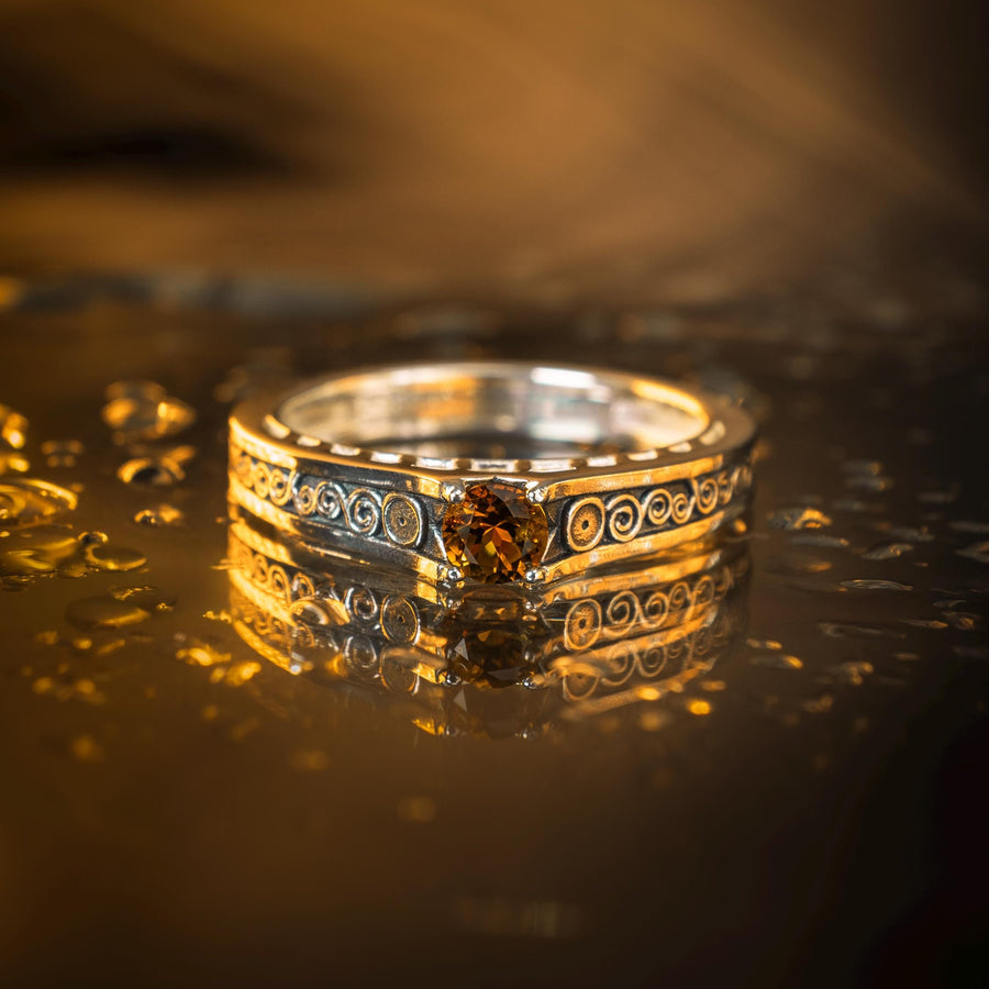 A detailed macro shot of a sterling silver engagement ring. The focal point is a round, honey-colored gemstone (Citrine or Madeira Topaz) set flush into the band. The shank is decorated with a continuous relief pattern of spirals and circles, mimicking the decorative robes found in Gustav Klimt's "Golden Phase" paintings. The silver features an oxidized finish, creating a dark background that makes the polished spirals pop. (etsy image id: 7142785932)