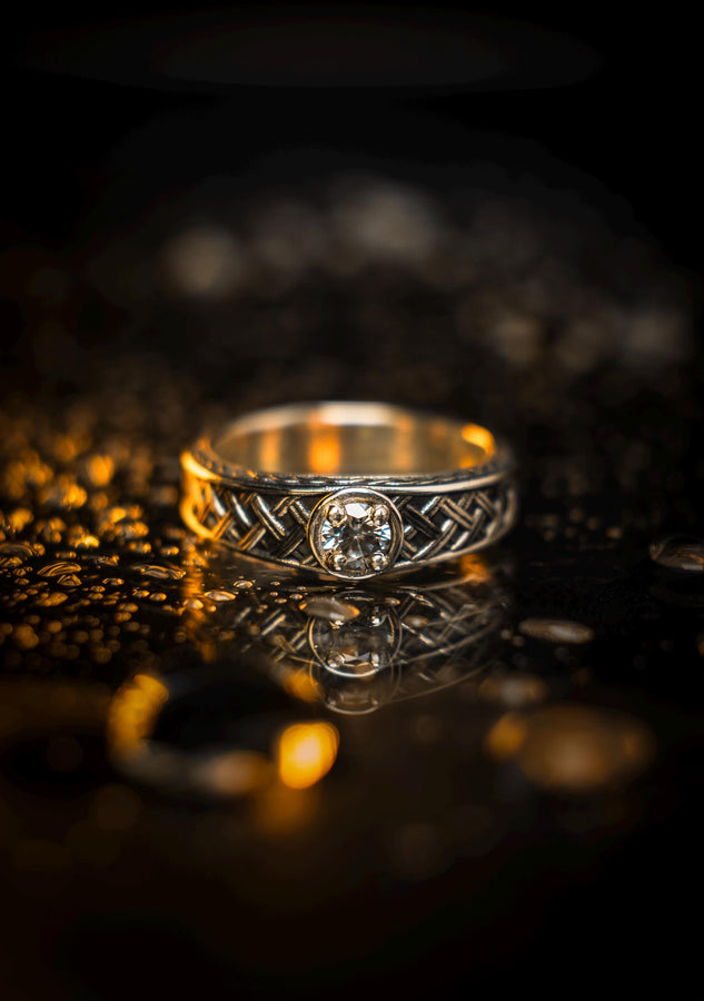 A warm, atmospheric shot of a matching wedding ring set. The engagement ring stands upright, showcasing a round white gemstone in a bezel setting. Lying flat beneath it is a matching wide band without a stone. Both rings share a cohesive design: a central diagonal "basket weave" pattern flanked by twisted rope borders. The silver is oxidized to reveal the intricate texture of the weave.