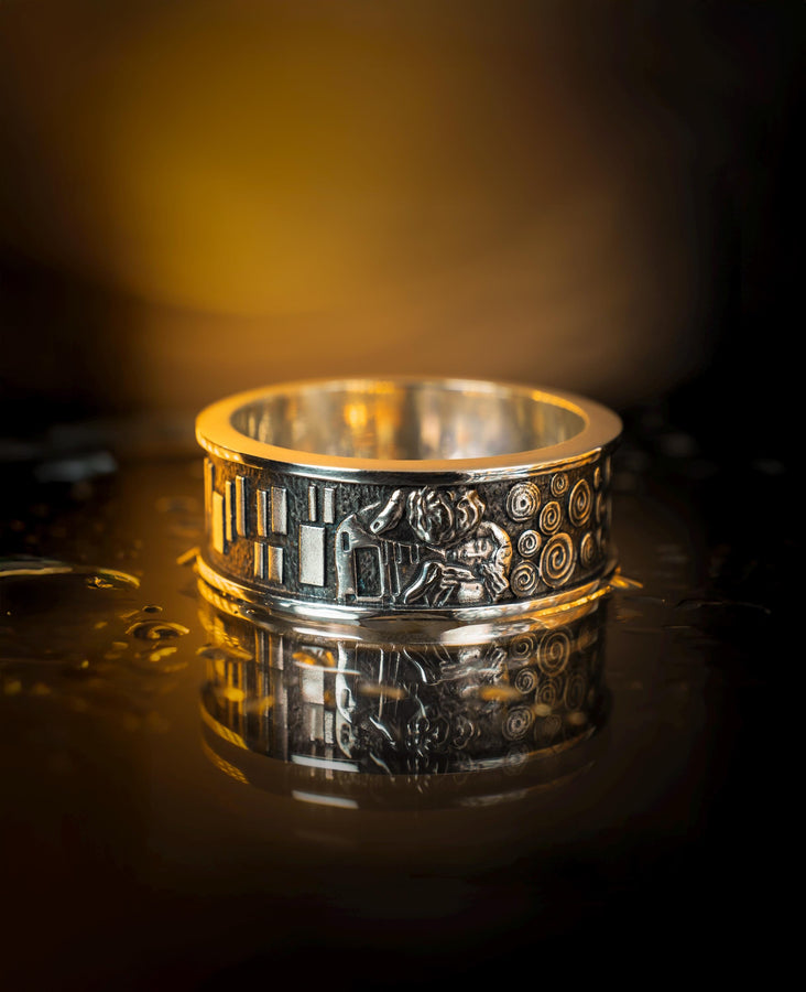 A cinematic studio shot of a matching two-piece ring set. The bottom ring is a wide silver band featuring a detailed relief carving of the couple from Gustav Klimt's "The Kiss." The top ring is a narrower engagement band that mimics the geometric patterns of the painting and holds a round, amber-colored gemstone. The background is a warm, golden haze, reflecting the "Golden Phase" of the original artwork.