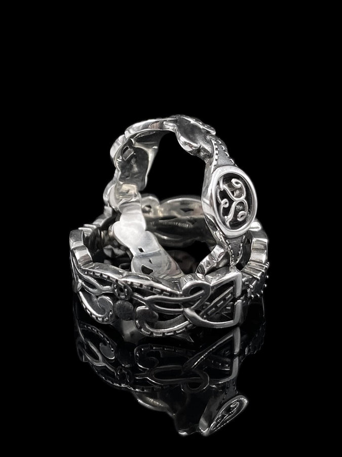A cinematic macro shot of a sterling silver ring bathed in warm, golden light. The focal point is the central oval face, which encloses a raised relief design of two spirals meeting in the center to suggest a heart shape. The band shoulders feature textured knotwork that leads up to the centerpiece. The ring rests on a wet, reflective surface, creating a mirror image of the love knot below. (etsy image id: 7490345549)