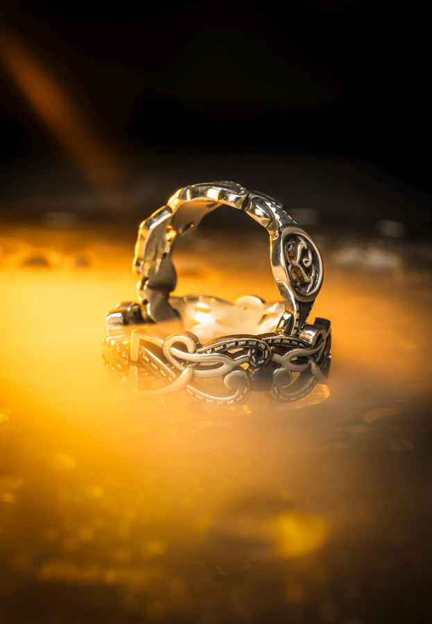 A cinematic close-up of a sterling silver ring lying on a reflective surface. The band features an "openwork" design, meaning the Celtic knot pattern is cut out, allowing light to pass through the loops. The silver strands weave over and under in a continuous infinity motif. The background is dark, but the ring is illuminated by a warm, golden mist. (etsy image id: 7442429936)