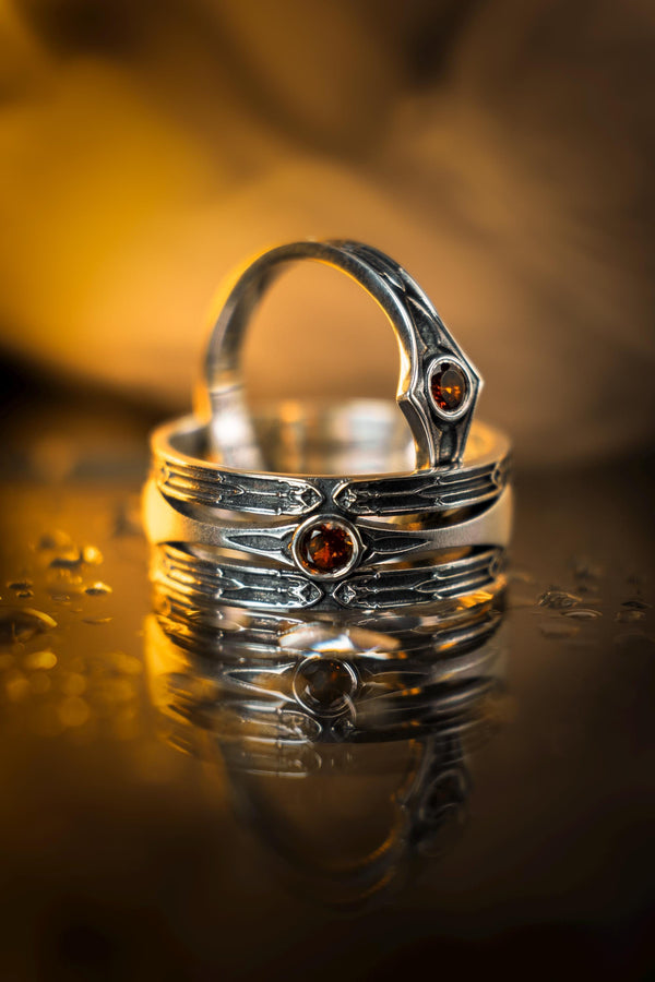 A detailed studio shot of a wide, heavy-gauge sterling silver ring. The focal point is a round, deep red gemstone (Garnet) bezel-set in the center. Flanking the stone are three parallel, tapered grooves that resemble the fletching of an arrow or the futuristic paneling of armor. The metal surfaces are brushed silver, while the deep grooves are blackened with oxidation to emphasize the directional lines. (etsy image id: 7441158070)