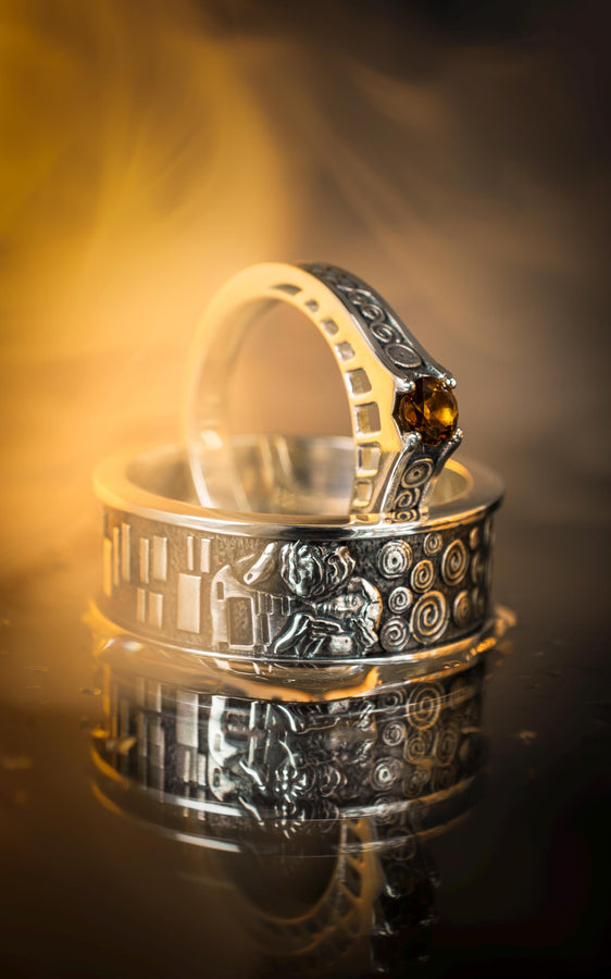 A cinematic studio shot of a matching two-piece ring set. The bottom ring is a wide silver band featuring a detailed relief carving of the couple from Gustav Klimt's "The Kiss." The top ring is a narrower engagement band that mimics the geometric patterns of the painting and holds a round, amber-colored gemstone. The background is a warm, golden haze, reflecting the "Golden Phase" of the original artwork.
