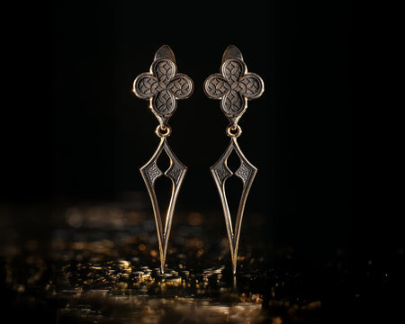 A clean studio shot of a pair of articulated sterling silver earrings resting on a matte grey surface. The top stud is a four-lobed "quatrefoil" shape filled with a detailed lattice texture of overlapping circles. The bottom part is a dangling, sharp "spear" or "arrowhead" shape with a geometric cutout in the center. The silver is oxidized, highlighting the texture in the stud and the sharp lines of the drop. (etsy image id: 7265852941)