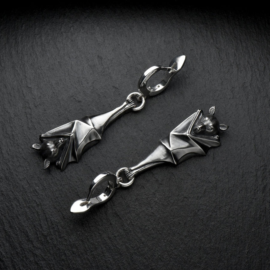 May include: A pair of silver-colored bat-shaped earrings. Each earring features a detailed bat design with outstretched wings and a small loop for attachment. The earrings are suspended from a curved hook. The background is a dark, textured surface. A detailed close-up studio shot of a pair of sterling silver dangle earrings shaped like flying foxes. Each earring features a highly textured sleeping fruit bat hanging upside down, wrapped securely (etsy image id: 7368202736)