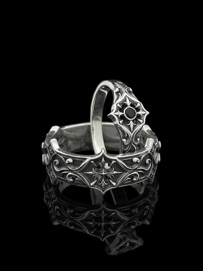 A cinematic studio shot of a matching two-piece ring set on dark velvet. The top ring stands upright, featuring a diamond-shaped face with a four-pointed star design and a small, round black stone in the center. The bottom ring lies flat, displaying a wide band with a repeating pattern of the same star/cross motif connected by swirling filigree. The silver is polished on the ridges and dark in the background.
