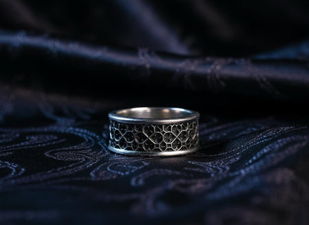A detailed macro shot of a wide sterling silver ring featuring a repeating "quatrefoil" design. The pattern consists of four-lobed geometric shapes that interlock like the stone tracery of a Gothic cathedral window. The raised silver lines are polished to a bright shine, while the recessed areas are chemically oxidized to a pitch-black matte finish. The contrast highlights the mathematical precision of the medieval-inspired lattice. (etsy image id: 7434448224)
