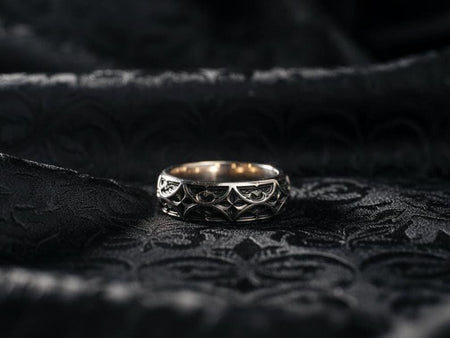 A studio profile shot of a wide sterling silver band standing upright on a black reflective surface. The ring features a continuous, deep-relief pattern of Gothic tracery—interlocking pointed arches and stylized four-petaled flowers. The raised lines are highly polished silver, while the recessed background is chemically oxidized to a matte black, creating a sharp, graphic contrast. (etsy image id: 7440812790)