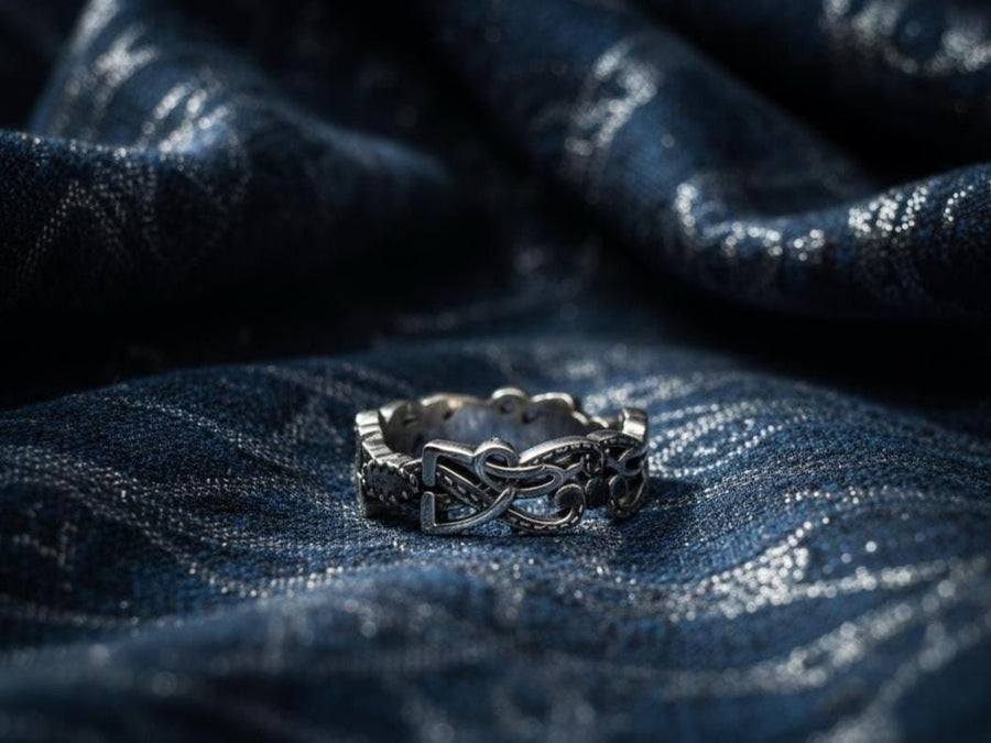 A cinematic close-up of a sterling silver ring lying on a reflective surface. The band features an "openwork" design, meaning the Celtic knot pattern is cut out, allowing light to pass through the loops. The silver strands weave over and under in a continuous infinity motif. The background is dark, but the ring is illuminated by a warm, golden mist. (etsy image id: 7442429936)