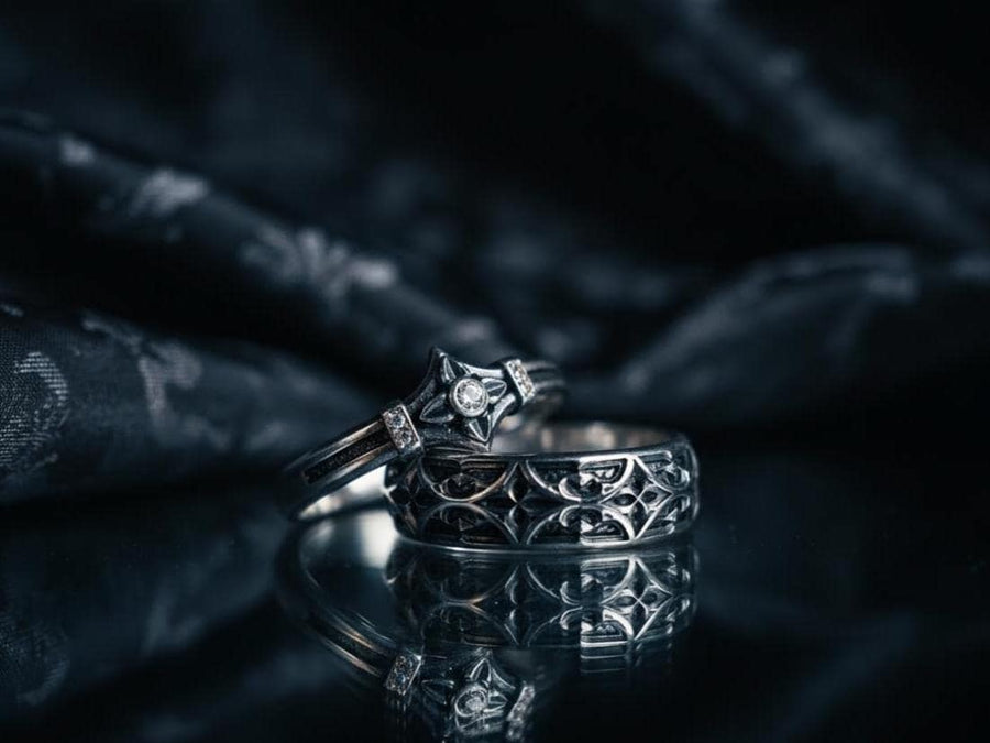 A high-resolution macro photograph of a matching sterling silver gothic wedding ring set resting on softly draped, luxurious white satin. The set features a wider masculine band and a slightly narrower feminine band. Both rings display a continuous engraved arcade of pointed medieval arches along the bottom edge, centering on a polished geometric rhombus frame that holds a brilliant round white gemstone. The polished silver beautifully contrasts  (etsy image id: 7928017042)