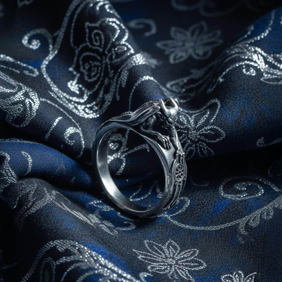 A detailed profile shot of a sculptural sterling silver ring. The band is formed by the body of a crouching gargoyle. The creature features distinct horns, a snarling face, and muscular limbs. Its bat-like wings fold back to form the sides of the ring shank. In its front claw, it holds a small, circular charm with a web pattern, resembling a dreamcatcher. The silver is heavily oxidized black in the crevices to accentuate the details. (etsy image id: 7497960785)