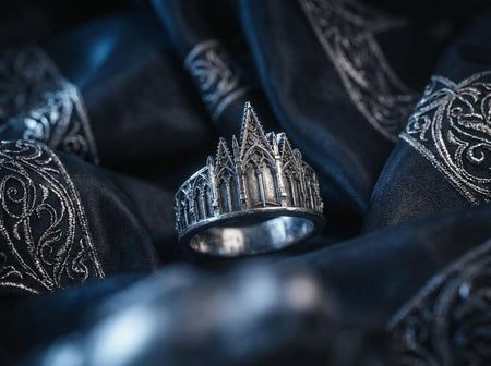 A clean studio shot of a sculptural sterling silver ring resting on a matte grey surface. The ring depicts a Gothic building facade with a tall, triangular central peak (gable) filled with geometric tracery. Flanking the center are lower pointed arches and vertical buttresses that wrap around the band. The silver is matte-finished with dark oxidation in the recesses to mimic stone shadows. (etsy image id: 7500126009)