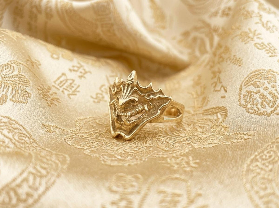 A close-up studio shot of a sterling silver ring featuring a detailed Japanese Oni mask. The demon face has sharp fangs, a wide menacing grin, and two distinct horns protruding from its forehead. The band features openwork cutouts on the sides, merging seamlessly with the mask. The metal is heavily oxidized to a dark charcoal in the recesses, making the highly polished silver ridges of the fierce expression pop against the dark reflective backgro (etsy image id: 7959798327)