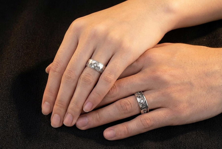 A high-resolution macro photograph of a matching sterling silver gothic wedding ring set resting on softly draped, luxurious white satin. The set features a wider masculine band and a slightly narrower feminine band. Both rings display a continuous engraved arcade of pointed medieval arches along the bottom edge, centering on a polished geometric rhombus frame that holds a brilliant round white gemstone. The polished silver beautifully contrasts  (etsy image id: 7928017042)