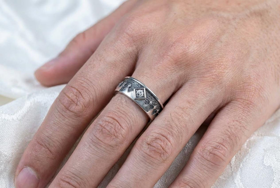A high-resolution macro photograph of a matching sterling silver gothic wedding ring set resting on softly draped, luxurious white satin. The set features a wider masculine band and a slightly narrower feminine band. Both rings display a continuous engraved arcade of pointed medieval arches along the bottom edge, centering on a polished geometric rhombus frame that holds a brilliant round white gemstone. The polished silver beautifully contrasts  (etsy image id: 7928017042)