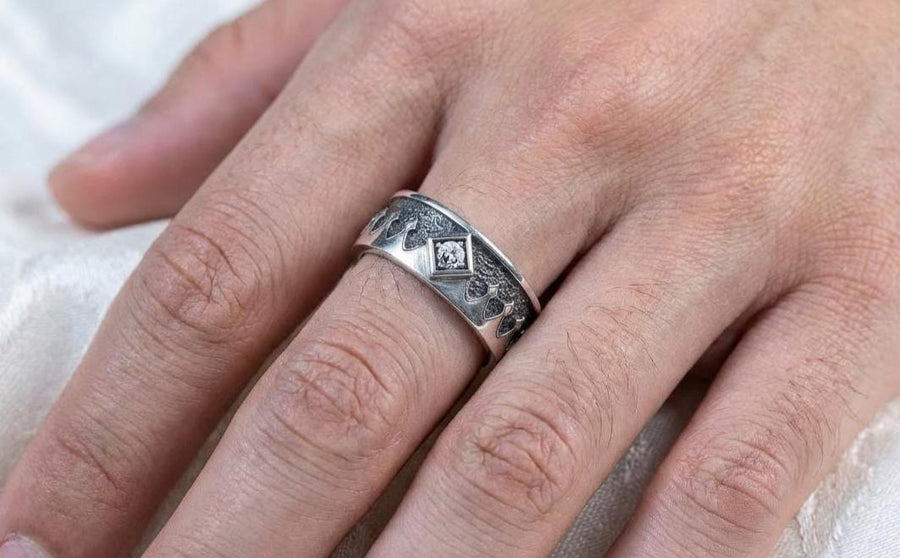 A high-resolution macro photograph of a matching sterling silver gothic wedding ring set resting on softly draped, luxurious white satin. The set features a wider masculine band and a slightly narrower feminine band. Both rings display a continuous engraved arcade of pointed medieval arches along the bottom edge, centering on a polished geometric rhombus frame that holds a brilliant round white gemstone. The polished silver beautifully contrasts  (etsy image id: 7928017042)