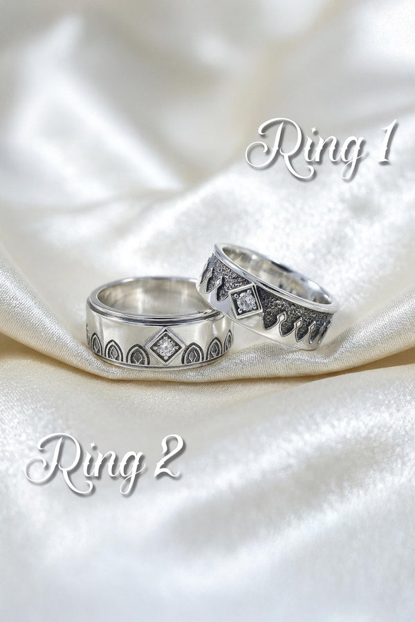 A high-resolution macro photograph of a matching sterling silver gothic wedding ring set resting on softly draped, luxurious white satin. The set features a wider masculine band and a slightly narrower feminine band. Both rings display a continuous engraved arcade of pointed medieval arches along the bottom edge, centering on a polished geometric rhombus frame that holds a brilliant round white gemstone. The polished silver beautifully contrasts  (etsy image id: 7928017042)