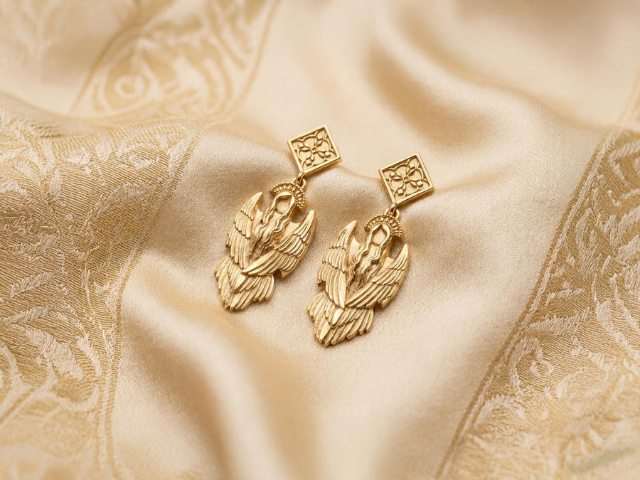 A detailed studio macro shot of a pair of articulated sterling silver earrings on a dark reflective surface. The top stud is a diamond shape enclosing a gothic floral cross. It suspends a highly detailed seraphim angel figure featuring a faceless head crowned with a studded halo. Her large, textured wings fold downwards into a feathered skirt. The silver is oxidized, leaving the recesses dark while the raised feathers gleam. (etsy image id: 8003029872)