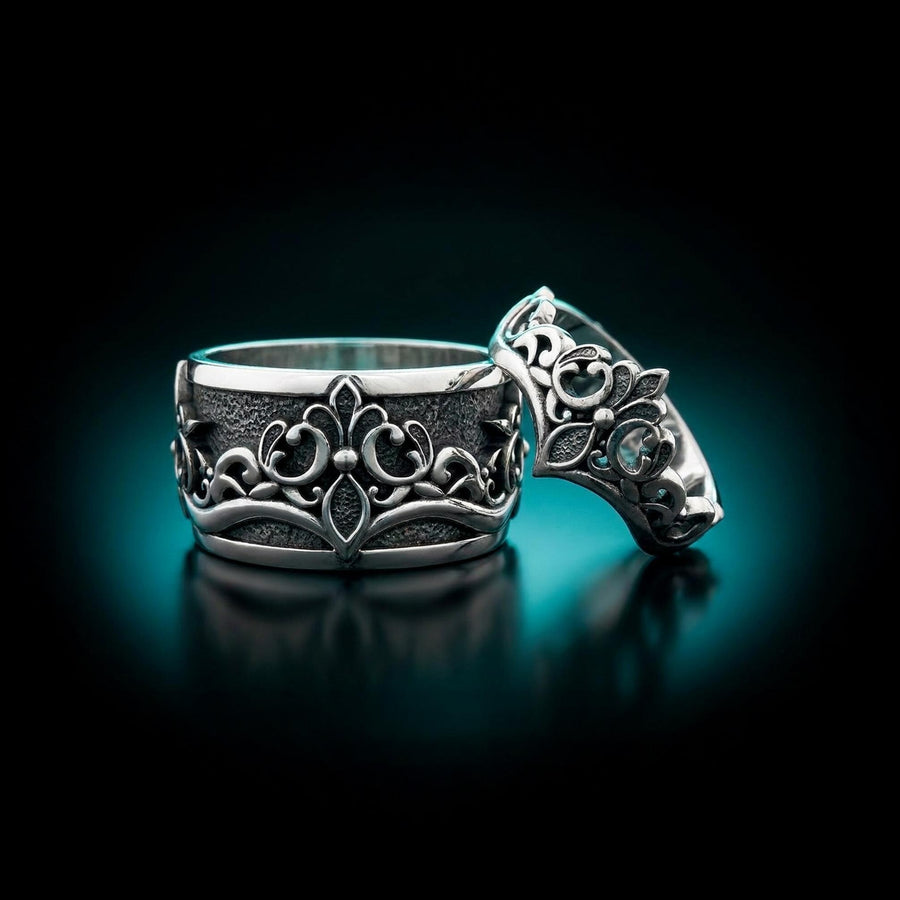 A close-up studio shot of a matching sterling silver gothic wedding ring set resting on a dark reflective surface. The set includes a wide band and a matching V-shaped chevron ring that nests perfectly against it. Both rings feature raised, polished silver scrollwork and fleur-de-lis motifs set against a deeply oxidized, stippled dark grey background. The intricate relief mimics medieval stone carvings, offering a rugged yet highly sophisticated
